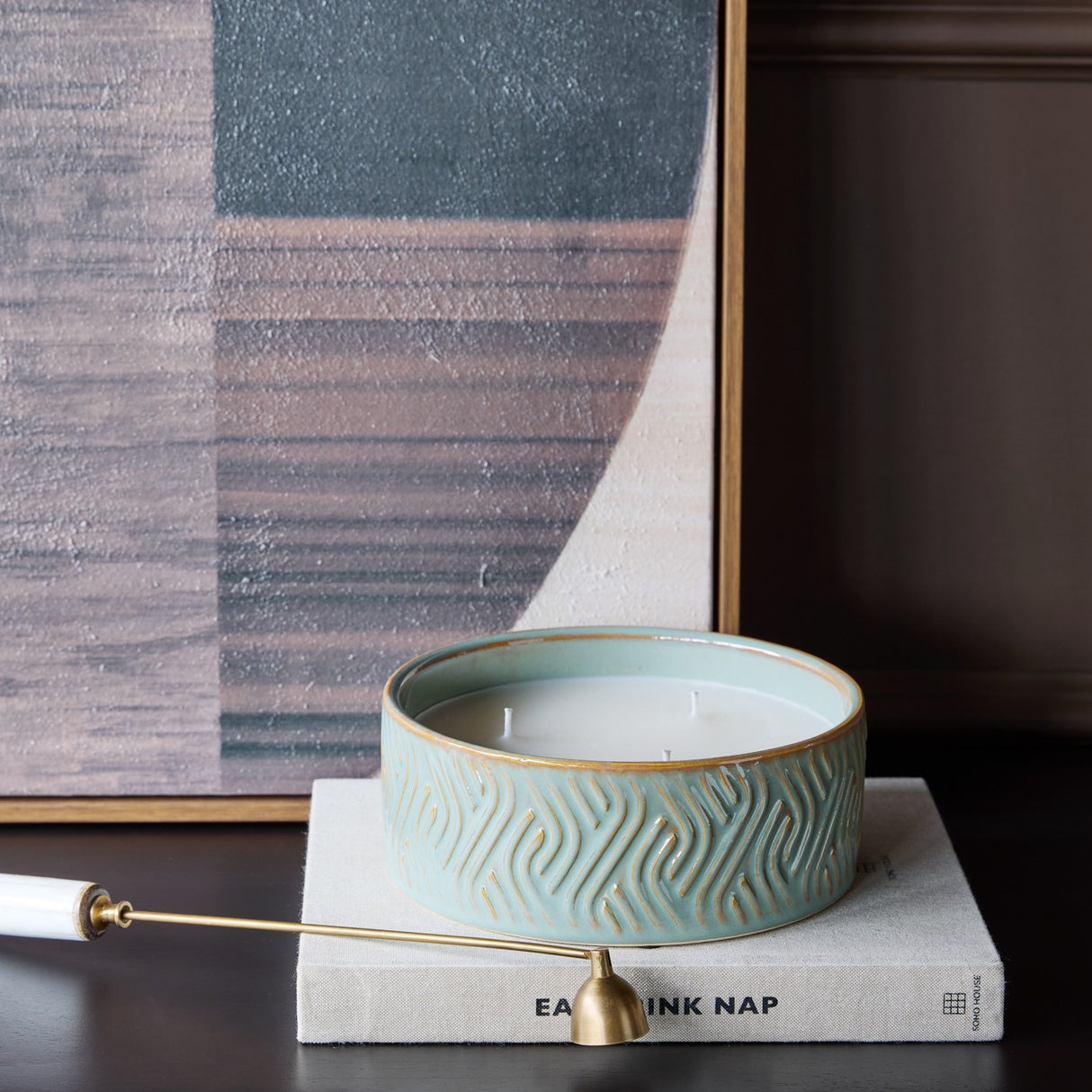 Libra Interiors embossed ceramic triple-wick candle in soft blue-green, resting on a linen-bound book beside a framed abstract artwork.