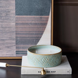Libra Interiors embossed ceramic triple-wick candle in soft blue-green, resting on a linen-bound book beside a framed abstract artwork.