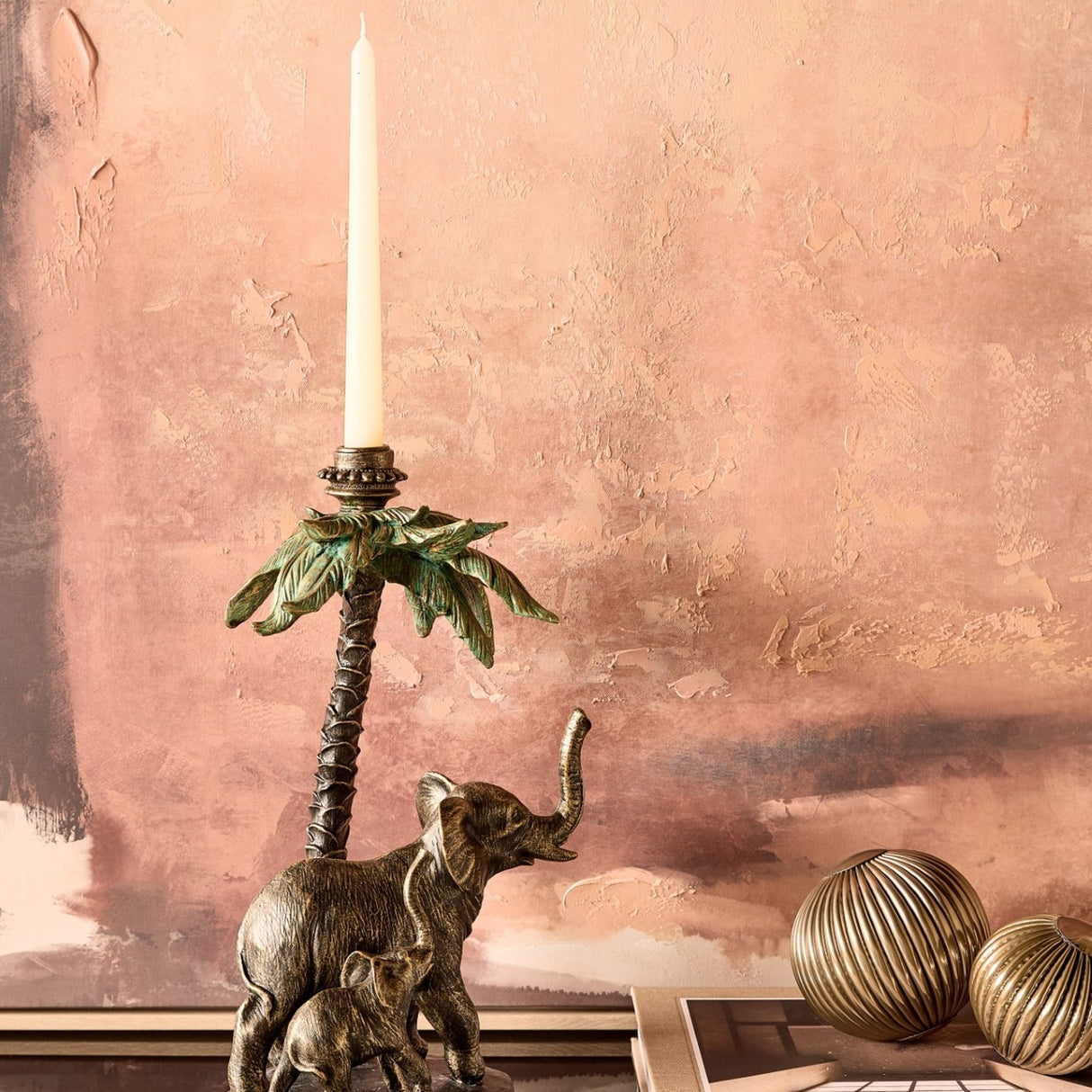 Libra Interiors Palm and Elephant resin candle holder in gold, with a mother and calf beneath a palm tree holding a taper candle, on a mantel.