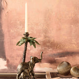 Libra Interiors Palm and Elephant resin candle holder in gold, with a mother and calf beneath a palm tree holding a taper candle, on a mantel.