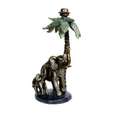 Libra Interiors Palm and Elephant resin candle holder in aged gold, showing a mother elephant and calf beneath a green-tinted palm tree taper fitting.