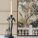 Libra Interiors Giraffe & Palm resin candle holder in gold with verdigris fronds, holding a cream taper beside a framed botanical print.