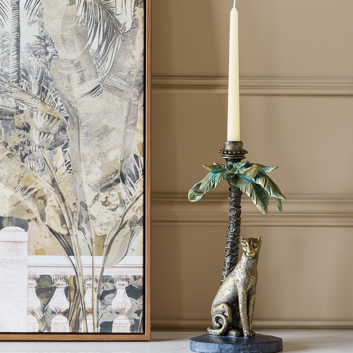 Libra Interiors Palm Tree and Leopard candle holder in gold resin with verdigris fronds, holding a cream taper on a console beside botanical wall art.
