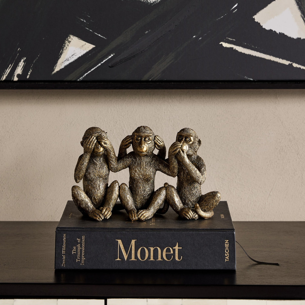 Libra Interiors Textured Antique Gold Three Wise Monkeys resin sculpture displayed on a Monet book atop a dark console beneath abstract black artwork.