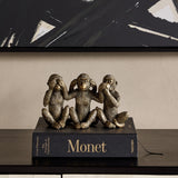 Libra Interiors Textured Antique Gold Three Wise Monkeys resin sculpture displayed on a Monet book atop a dark console beneath abstract black artwork.