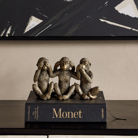 Libra Interiors Textured Antique Gold Three Wise Monkeys resin sculpture displayed on a Monet book atop a dark console beneath abstract black artwork.