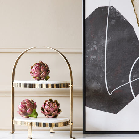 Libra Interiors Marble Two Tier Cake Stand in white with aged gold arched frame, holding artichokes beside a black and white abstract artwork.