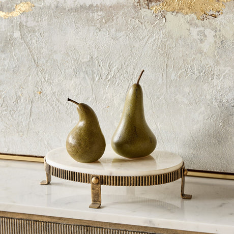 Libra Interiors Marble Oval Aged Gold Cake Stand with white marble top and ridged brass-toned base, styled with two olive green pears against a textured wall.
