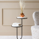 Libra Interiors Two-Tier Marble Top Bronze Side Table beside an armchair, with a book on the lower tier and an amber vase of dried grasses on top.