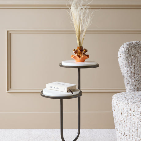 Libra Interiors Two-Tier Marble Top Bronze Side Table beside an armchair, with a book on the lower tier and an amber vase of dried grasses on top.