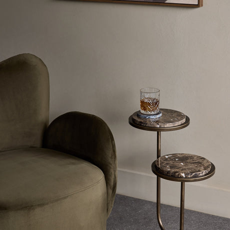 Libra Interiors two-tier Dark Emperador marble side table with staggered round tops on a bronze iron frame, beside an olive velvet armchair with a whisky glass.