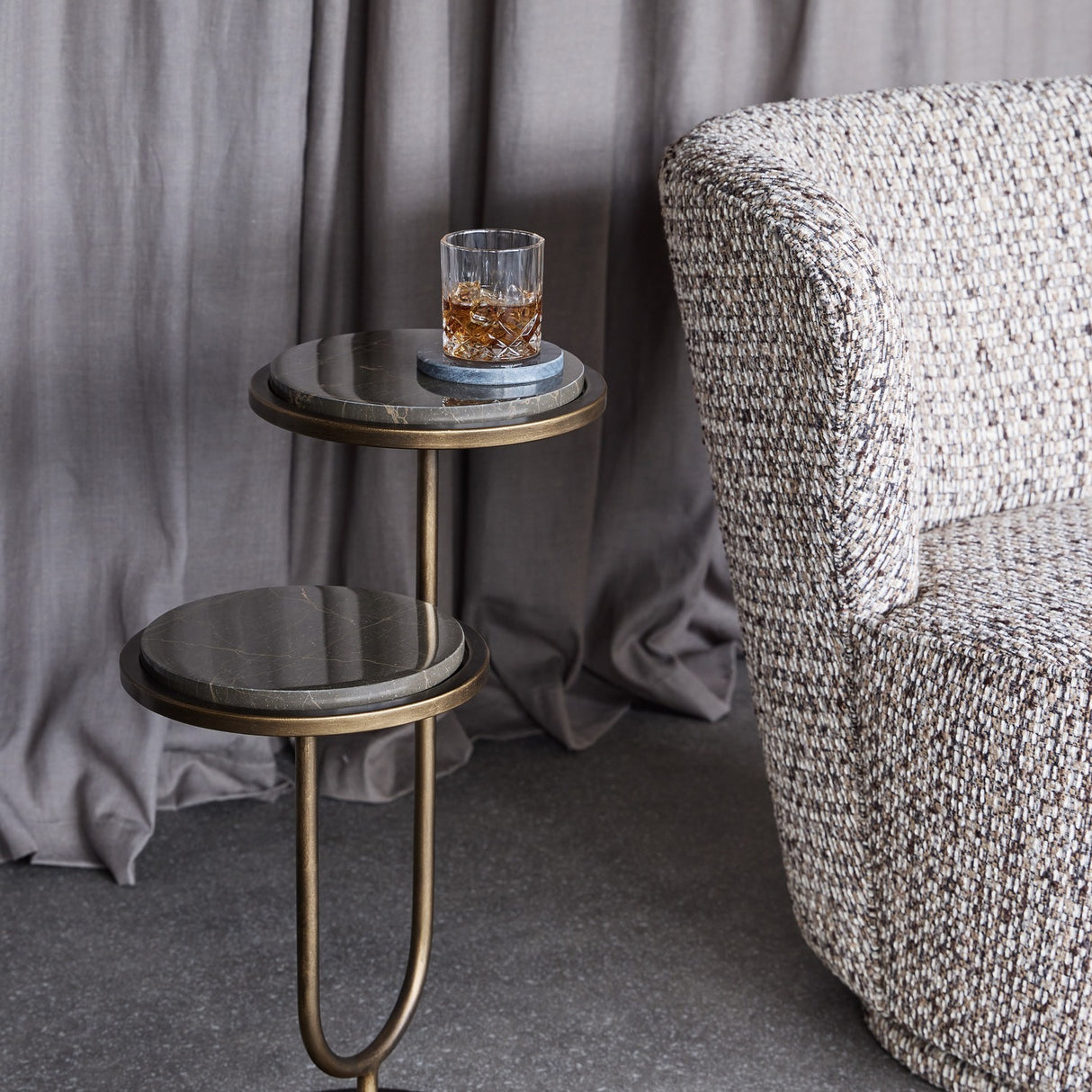 Libra Interiors Two-Tier Marble Burnished Gold Side Table in brown beside a tweed armchair, holding a crystal whisky glass on a coaster against grey curtains.