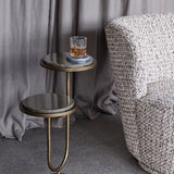 Libra Interiors Two-Tier Marble Burnished Gold Side Table in brown beside a tweed armchair, holding a crystal whisky glass on a coaster against grey curtains.
