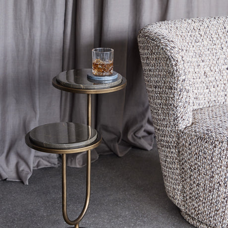 Libra Interiors Two-Tier Marble Burnished Gold Side Table in brown beside a tweed armchair, holding a crystal whisky glass on a coaster against grey curtains.