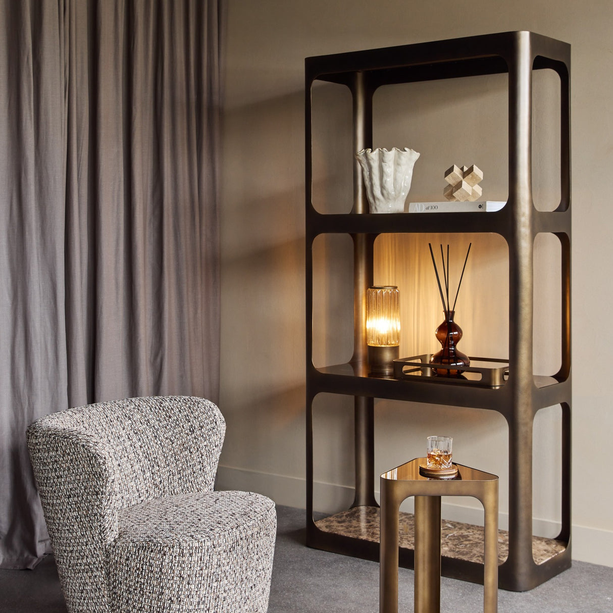 Libra Interiors Triangular Smoked Gold Iron Side Table in bronze with black tinted glass top, holding a whisky glass beside a bronze shelving unit and armchair.