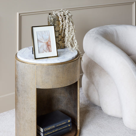 Libra Interiors Marble Top Bedside Table in gold with ridged metal frame, white marble top and tinted glass shelf, styled beside a cream chair.
