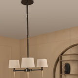 Libra Interiors 3-Light Linen Shade Bronze Chandelier with pale linen shades and gold tapered feet, hanging from an adjustable drop in a warm neutral room.