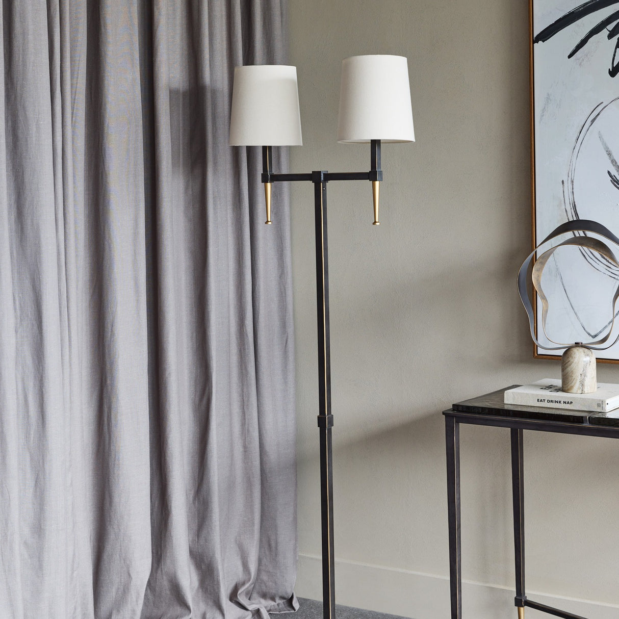 Libra Interiors Twin Shade Bronze Iron Floor Lamp with two bleached linen shades and gilded accents, styled beside grey curtains and a black console table.