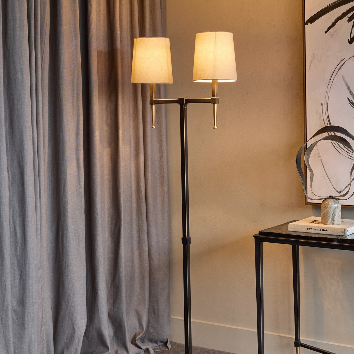 Libra Interiors Twin Shade Bronze Iron Floor Lamp with two bleached linen shades and gold accents, beside grey curtains and a black console table.