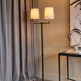 Libra Interiors Twin Shade Bronze Iron Floor Lamp with two bleached linen shades and gold accents, beside grey curtains and a black console table.