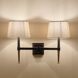 Libra Interiors Twin Linen Shade Bronze Wall Light with dark iron frame, gold tapered feet, square backplate and two bleached linen shades illuminated.
