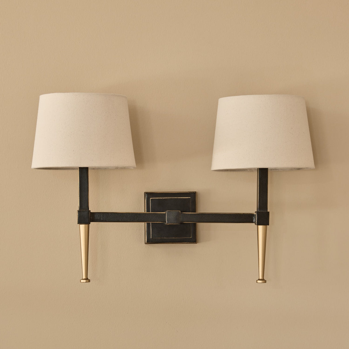 Libra Interiors Twin Linen Shade Bronze Wall Light with dark iron frame, brushed gold tapered arms, square backplate and two bleached linen shades.