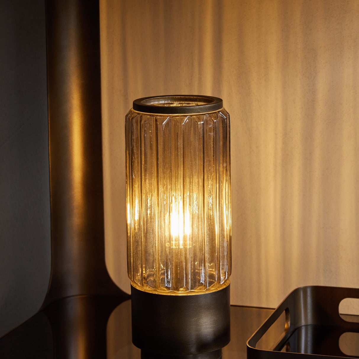 Libra Interiors Fluted Glass Uplighter Table Lamp in brown gold, lit on a dark surface, showing ribbed cylindrical shade glowing above a bronze metal base.