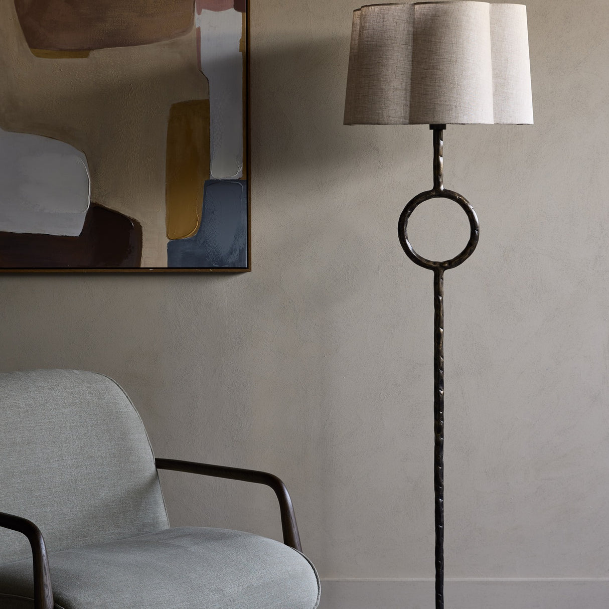 Libra Interiors Hammered Iron floor lamp in bronze with circular stem detail and scalloped linen shade, beside a grey armchair and abstract artwork.