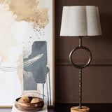 Libra Interiors Hammered Iron Ring Table Lamp in bronze with scalloped linen shade and marble base, styled beside abstract art and a wooden bowl.