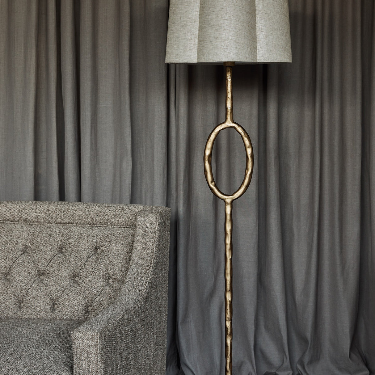 Libra Interiors Hammered Oval Gold Floor Lamp with iron stem, open oval detail and scalloped ivory linen shade beside a grey tufted armchair and curtains.