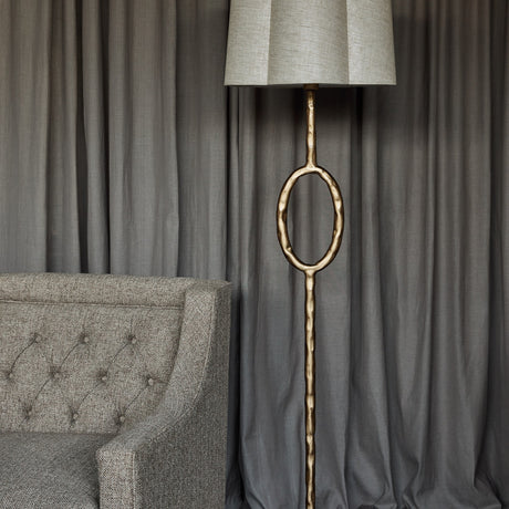 Libra Interiors Hammered Oval Gold Floor Lamp with iron stem, open oval detail and scalloped ivory linen shade beside a grey tufted armchair and curtains.