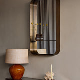 Libra Interiors Smoked Gold Iron Shelf Wall Mirror mounted in a hallway, with two small ceramic vases of yellow craspedia on its integrated shelf.