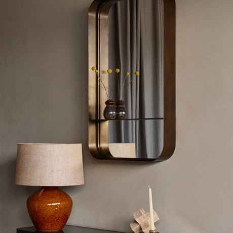 Libra Interiors Smoked Gold Iron Shelf Wall Mirror mounted in a hallway, with two small ceramic vases of yellow craspedia on its integrated shelf.