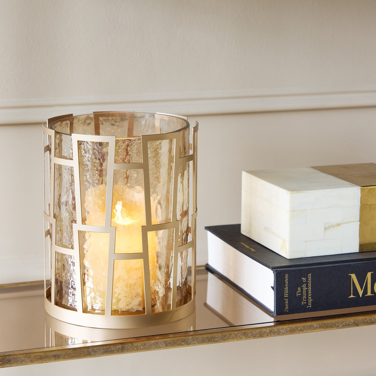 Libra Interiors Hammered Glass Geometric Gold Lantern with lit pillar candle, displayed on a mirrored console beside stacked books and a bone box.