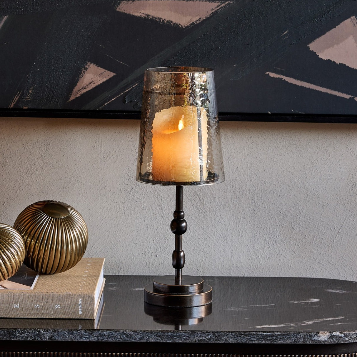 Libra Interiors Hammered Glass Candle Lantern in bronze with tapered copper-brown shade on turned iron base, lit on a dark console beside brass ornaments.