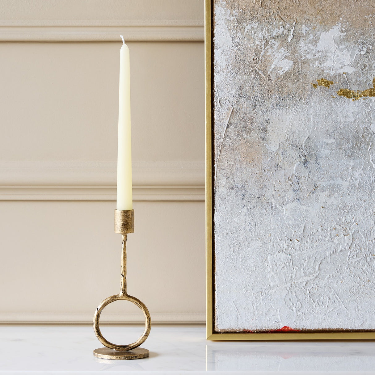 Libra Interiors Ring Iron Antique Gold Candle Holder with sculptural ring base and ivory taper candle, styled beside a textured silver and gold artwork.