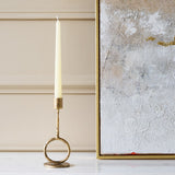 Libra Interiors Ring Iron Antique Gold Candle Holder with sculptural ring base and ivory taper candle, styled beside a textured silver and gold artwork.