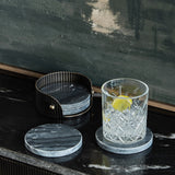 Libra Interiors Marble Round Set of 4 Coasters in black with veining, shown in a ribbed dark bronze holder beside a cut glass tumbler on a dark surface.