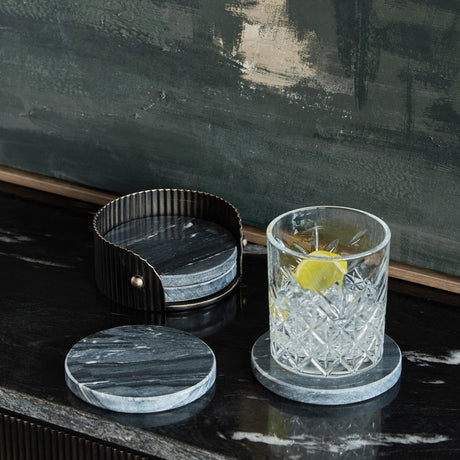 Libra Interiors Marble Round Set of 4 Coasters in black with veining, shown in a ribbed dark bronze holder beside a cut glass tumbler on a dark surface.