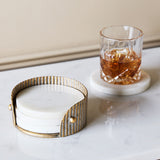 Libra Interiors Marble and Aged Gold Round Coaster set in a corrugated champagne gold iron holder beside a whisky glass on a marble coaster.