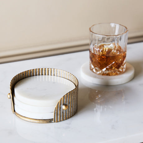 Libra Interiors Marble and Aged Gold Round Coaster set in a corrugated champagne gold iron holder beside a whisky glass on a marble coaster.