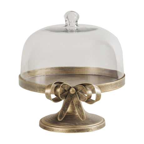 Libra Interiors Antiqued Gold Bow Glass Dome Cake Stand with iron pedestal, sculpted bow detail and lift-off glass cloche, on a plain white background.