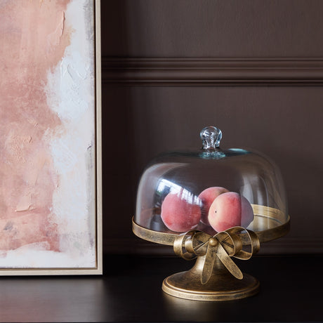 Libra Interiors Antiqued Gold Bow Glass Dome Cake Stand holding peaches on a footed pedestal with bow detail, beside a pink artwork on dark wall.