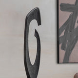 Libra Interiors textured cast aluminium abstract sculpture in matt black, with a sweeping hooked curve and open centre, beside a framed abstract artwork.