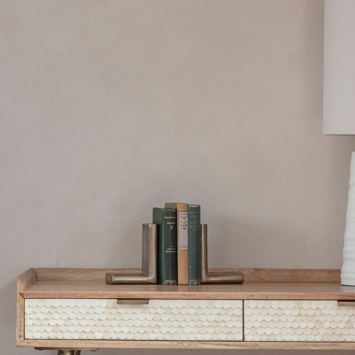 Libra Interiors ribbed cast aluminium L-shaped bookends in antique brass, holding vintage green hardbacks on a wooden console with scalloped bone drawers.