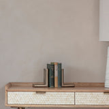 Libra Interiors ribbed cast aluminium L-shaped bookends in antique brass, holding vintage green hardbacks on a wooden console with scalloped bone drawers.