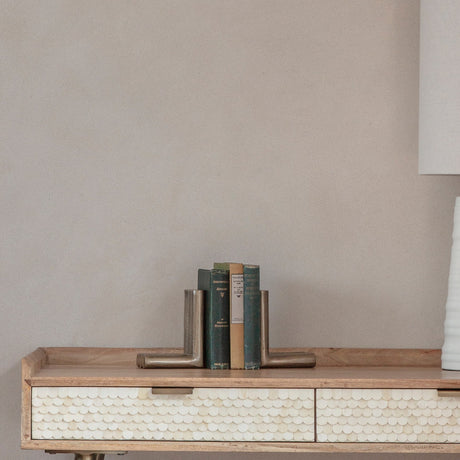 Libra Interiors ribbed cast aluminium L-shaped bookends in antique brass, holding vintage green hardbacks on a wooden console with scalloped bone drawers.