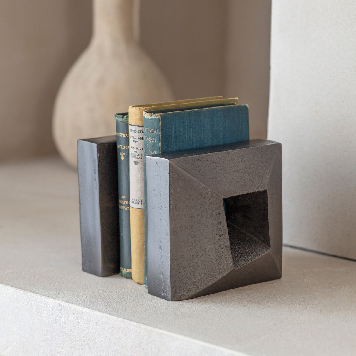 Libra Interiors Textured Cast Aluminium Bookends in grey holding vintage cloth-bound books, with geometric square form and hollowed centre on a pale shelf.