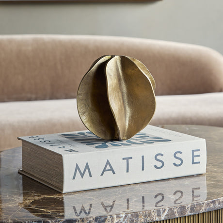 Libra Interiors Cast Aluminium Petal Sculpture in antique brass, displayed on a Matisse book atop a marble table with a blush velvet sofa behind.