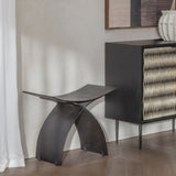 Libra Interiors Cast Aluminium Curved Stool in black with arched legs and concave seat, styled in a hallway beside a woven-front cabinet.